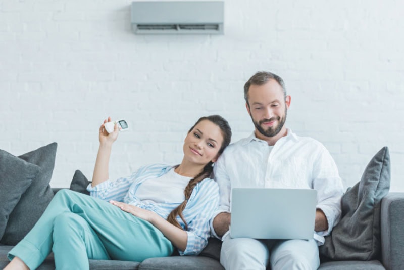 couple sitting on couch, using a remote to adjust their ductless ac unit.